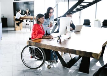 Hispanic transgender woman sitting in wheelchair and coworker looking at computer at workplace in Latin America