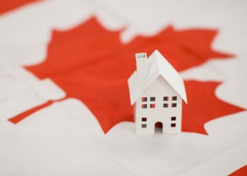 A small white miniature house sits on the Canadian flag, illustrating the concept of immigrating to Canada and living there.