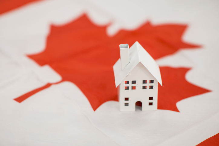 A small white miniature house sits on the Canadian flag, illustrating the concept of immigrating to Canada and living there.