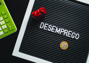 The word unemployment written in Brazilian portuguese in a frame with letters. In the composition of the image: an alarm clock, a calculator and a Brazilian Real coin.