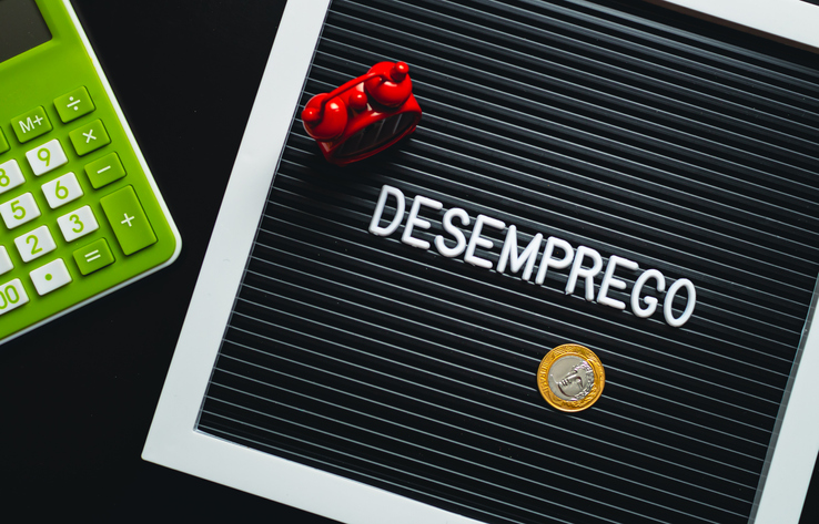 The word unemployment written in Brazilian portuguese in a frame with letters. In the composition of the image: an alarm clock, a calculator and a Brazilian Real coin.