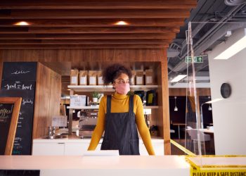 Female Small Business Owner Of Coffee Shop In Mask Standing Behind Counter During Health Pandemic