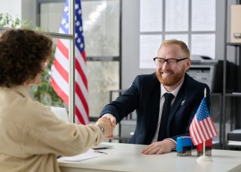 Portrait of smiling male consultant shaking hands with client over table in US Immigration office