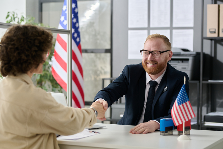 Portrait of smiling male consultant shaking hands with client over table in US Immigration office