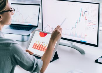 Crop back view of modern woman accounting statistical data while exploring charts on monitor of computer working at desk in office