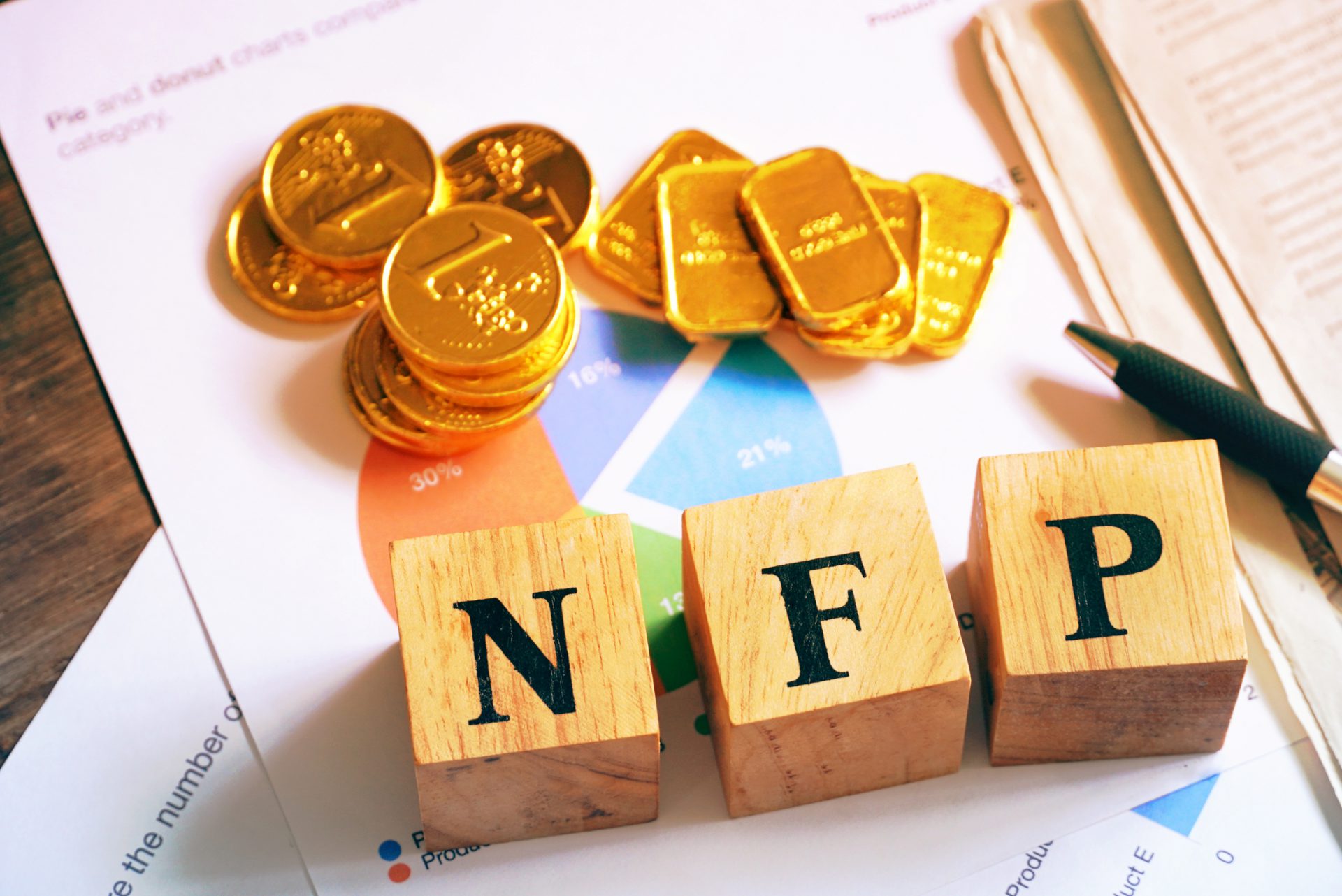 Text "NFP" on wood cube with gold bar and newspaper on the table, economic data concept
