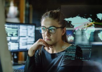 Stock image of a young woman, wearing glasses, surrounded by computer monitors in a dark office. In front of her there is a see-through displaying showing a map of the world with some data.