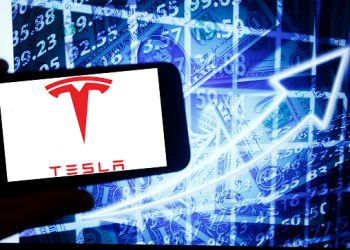 The logo of the Tesla Inc is seen on a screen of a smartphone next to a screen with an illustration ofthe stock market. Tesla is listed in Nasdaq. The Nasdaq is the second-largest stock exchange in the world after the New York Stock Exchange. (Photo by Alexander Pohl/NurPhoto via Getty Images)