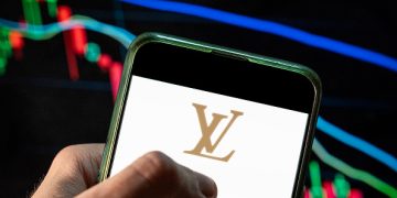 CHINA - 2021/12/09: In this photo illustration the French luxury fashion brand Louis Vuitton (LV) logo seen displayed on a smartphone with an economic stock exchange index graph in the background. (Photo Illustration by Budrul Chukrut/SOPA Images/LightRocket via Getty Images)